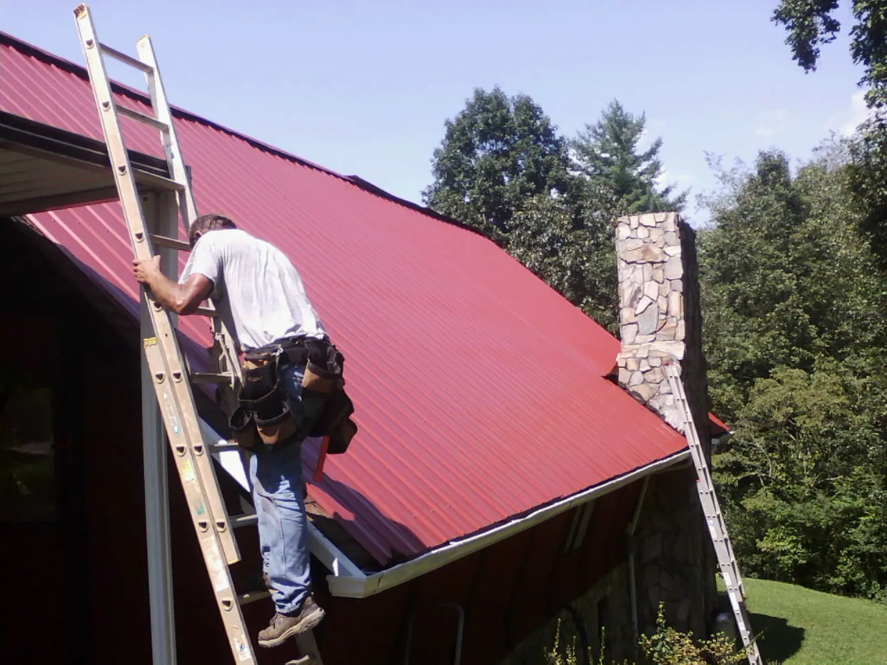 Roofer on a ladder beside a red metal roof and stone chimney — Soffit & Fascia Repair by Roofing Brothers in Perth Amboy